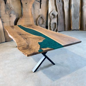 May include: A large wooden dining table with a live edge top and a green epoxy river running through the center. The table is supported by a black metal X-shaped base.