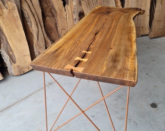 Può includere: Un tavolo in legno con un piano a bordo vivo e una base in metallo rame. Il piano del tavolo ha una finitura in legno naturale e presenta un'unica venatura. La base è realizzata con sottili aste di metallo angolate che creano un design geometrico.