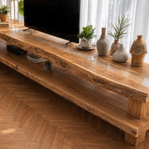 Può includere: Un mobile TV in legno a due livelli con un design naturale a bordo vivo. Il mobile è realizzato in legno marrone chiaro e presenta un ripiano inferiore. Vasi decorativi e piante adornano la superficie superiore. Il mobile è lungo circa 183 cm.