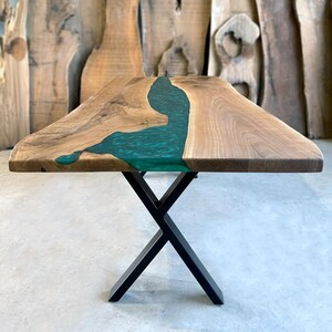 May include: A wooden dining table with a live edge top and a green epoxy river running through the center. The table is supported by a black metal X-shaped base.