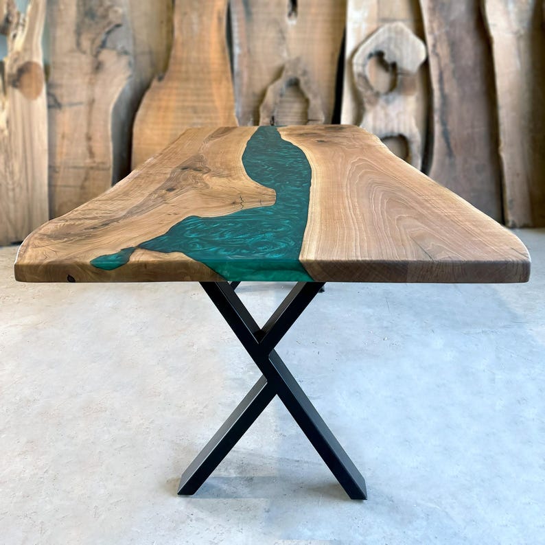 May include: A wooden dining table with a live edge top and a green epoxy river running through the center. The table is supported by a black metal X-shaped base.