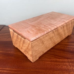May include: A rectangular wooden box featuring a light-colored, wavy-grained lid that contrasts with the darker, textured wood of the base. The box has a smooth finish and a slightly tapered design with small, carved feet at the bottom corners.