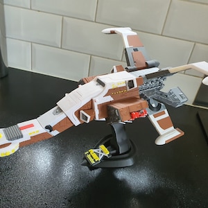 May include: A white and brown model spaceship with a long, thin body and two wings. The spaceship is on a black stand.