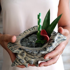 Puede incluir: Una taza de color gris en forma de concha marina llena de hielo y una ramita de menta. Dos pajitas rayadas verdes y una flor roja adornan la bebida.