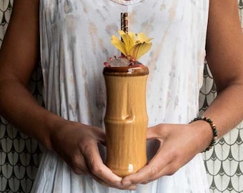 May include: A person holding a tall, brown ceramic tiki mug with a yellow flower garnish and a brown straw. The mug has a bamboo-like design.