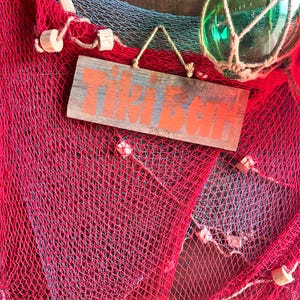 Può includere: Una rete da pesca rossa e blu con un cartello di legno che dice "Fish Bar" appeso ad essa. Il cartello è dipinto con lettere rosse e bianche.