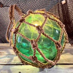 Pode incluir: Uma boia decorativa de vidro verde, envolta em uma rede de corda marrom. A esfera de vidro tem um brilho amarelo quente que emana de dentro. A rede de corda é amarrada ao redor da esfera e uma alça de corda é presa no topo.
