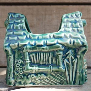 A green ceramic napkin holder shaped like a small house with a thatched roof and a window. The house has a textured surface and is decorated with details like a chimney, a door, and a fence.