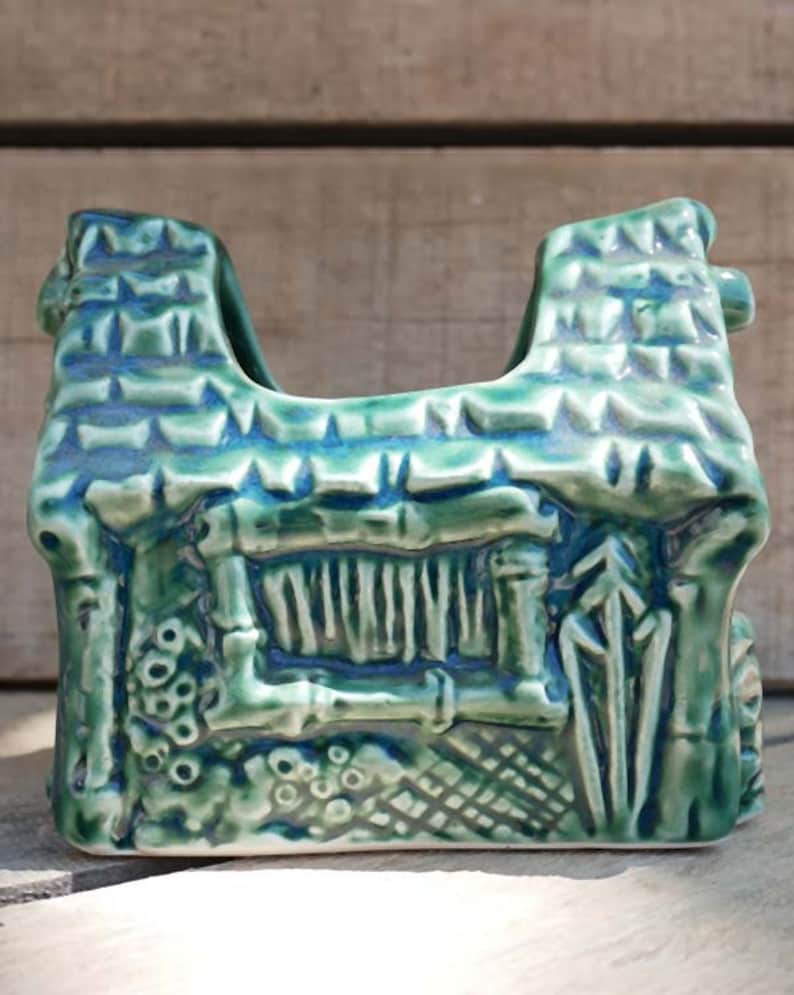 A green ceramic napkin holder shaped like a small house with a thatched roof and a window. The house has a textured surface and is decorated with details like a chimney, a door, and a fence.