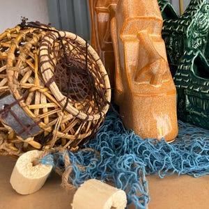 Könnte beinhalten: Eine Sammlung dekorativer Tiki-Artikel. Enthält einen geflochtenen Korb mit Netz, zwei hellfarbige Holzschwimmer, ein blaues Fischernetz und mehrere Tiki-Statuen aus Keramik in Orange- und Grüntönen.