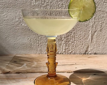 May include: A clear glass margarita glass with a yellow-colored drink and a lime wedge garnish. The glass has a brown, decorative stem with a tiki-style design.