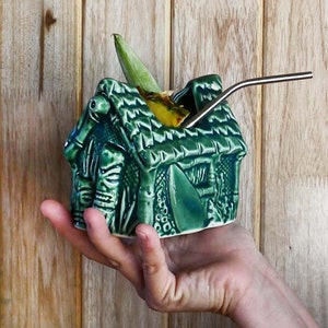 A green ceramic tiki mug shaped like a house with a thatched roof. The mug is filled with a yellow beverage and has a stainless steel straw sticking out of the top.