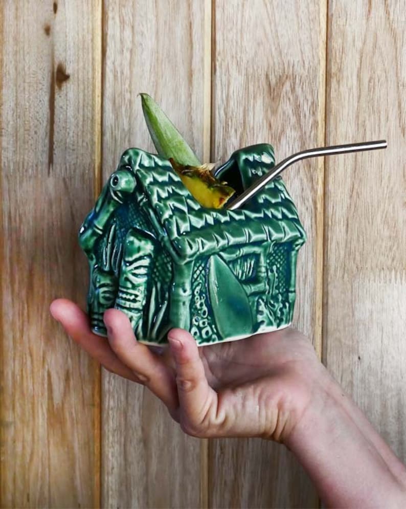 A green ceramic tiki mug shaped like a house with a thatched roof. The mug is filled with a yellow beverage and has a stainless steel straw sticking out of the top.