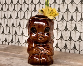 May include: A brown ceramic tiki mug shaped like a person with a flower on top. The mug is sitting on a wooden surface.