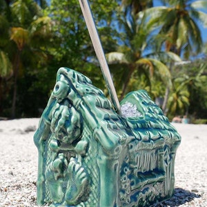 A green ceramic tiki mug shaped like a hut with a thatched roof. The mug has a straw sticking out of the top and is sitting on a white sandy beach.