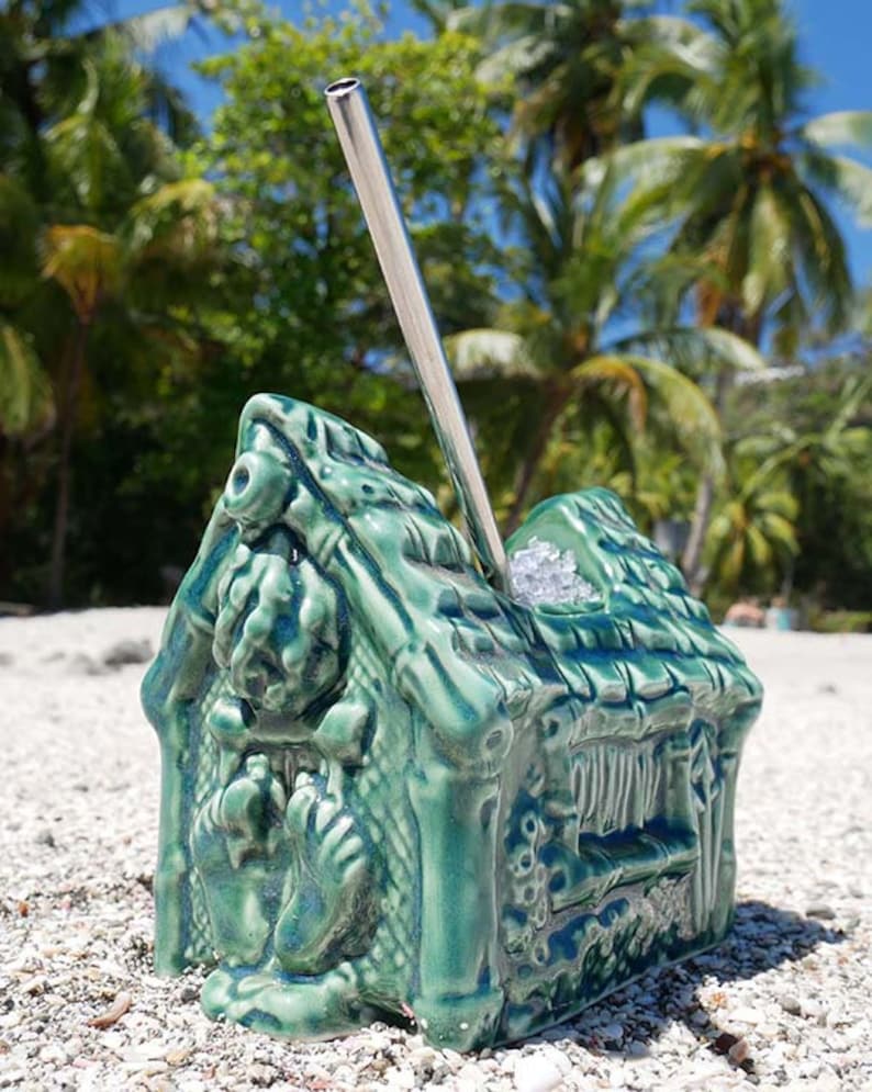 A green ceramic tiki mug shaped like a hut with a thatched roof. The mug has a straw sticking out of the top and is sitting on a white sandy beach.