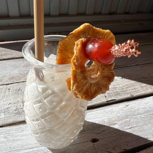 Könnte beinhalten: Ein klares Cocktailglas in Ananasform, gefüllt mit Eis und einem cremigen weißen Getränk. Garniert mit getrockneten Ananasscheiben, einer roten Kirsche und einem dekorativen kupferfarbenen Ananas-Pick. Ein Holzstrohhalm befindet sich im Glas.