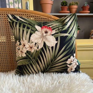 May include: Square decorative pillow with a tropical floral design. The pillow features a black background with large white and red flowers, and green palm leaves. The pillow is placed on a white fluffy surface.