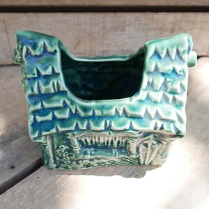 A green ceramic planter shaped like a house with a pointed roof. The planter has a textured surface and a small opening at the top for planting.