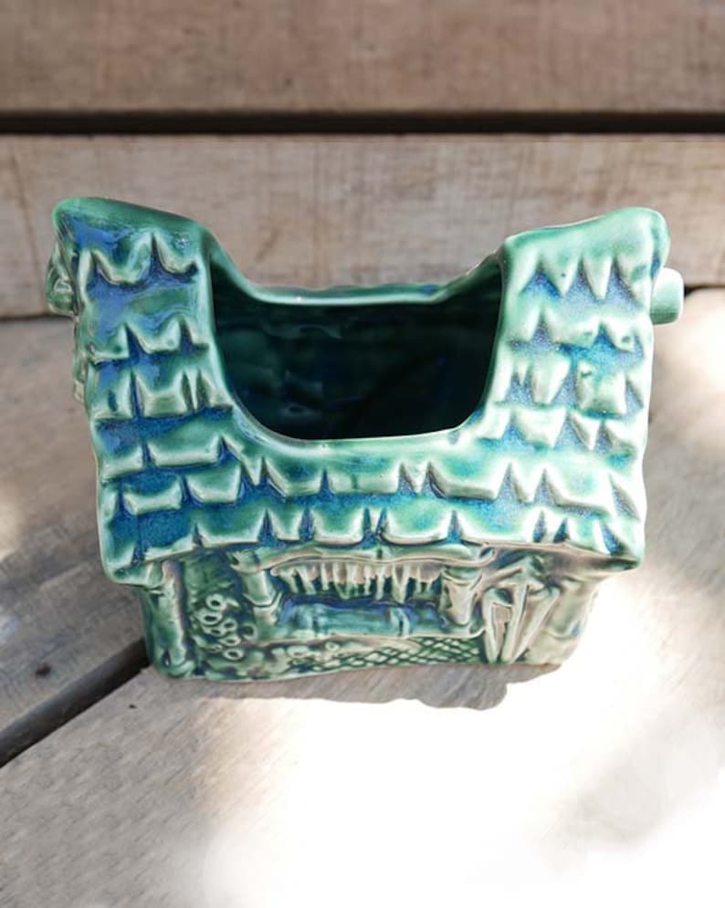 A green ceramic planter shaped like a house with a pointed roof. The planter has a textured surface and a small opening at the top for planting.