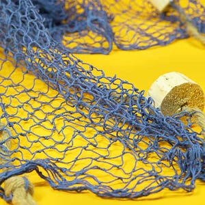 May include: A close-up of a blue decorative fishing net against a yellow backdrop. The net is made of knotted rope and has a textured appearance. Wooden floats are attached to the net with rope.