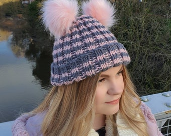 Puede incluir: Un gorro de punto gris y rosa con dos grandes pompones de piel sintética rosa. El gorro tiene un borde acanalado y un patrón tejido en gris y rosa. El gorro es un accesorio de invierno.