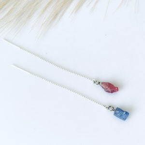 May include: A pair of silver chain earrings with a pink and a blue gemstone pendant. The earrings are delicate and simple.