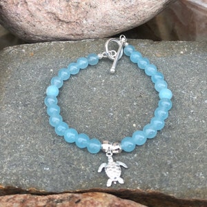 Könnte beinhalten: Ein hellblaues Perlenarmband mit einem silbernen Schildkrötenanhänger und einem silbernen Schiebeverschluss.