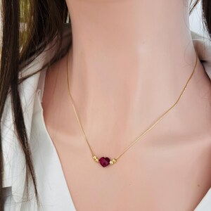 May include: A delicate gold necklace featuring a heart-shaped, faceted, deep red gemstone pendant. The pendant is flanked by two small gold beads. The necklace is worn against a white shirt.