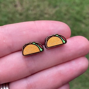 May include: A pair of wooden earrings shaped like tacos. The tacos are yellow with green lettuce and red meat fillings. The earrings have a glittery finish.