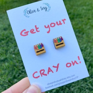 K&ouml;nnte beinhalten: Eine wei&szlig;e Karte mit dem Text "Olive & Ivy" und "Get your Cray on!"  Zwei kleine Holzohrringe in Form einer Schachtel mit Buntstiften sind an der Karte befestigt.