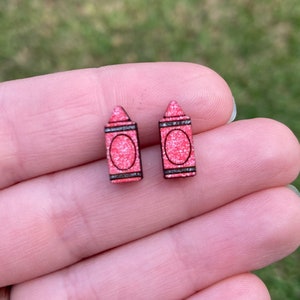 K&ouml;nnte beinhalten: Ein Paar rosa Glitzer-Kreide-Ohrstecker. Die Ohrringe sind aus Holz und haben einen schwarzen Umriss.