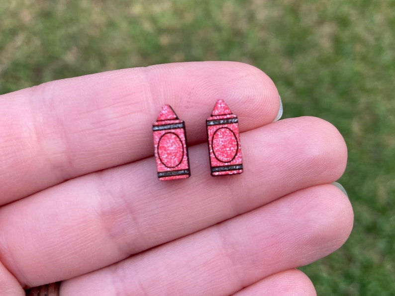 K&ouml;nnte beinhalten: Ein Paar rosa Glitzer-Kreide-Ohrstecker. Die Ohrringe sind aus Holz und haben einen schwarzen Umriss.