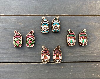May include: Set of six wooden gnome earrings with various designs. The gnomes are wearing red, green, and blue hats with white stripes. Some gnomes are holding a snowflake, a Christmas tree, and a gift.