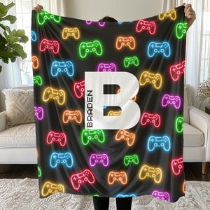 May include: Black blanket with a pattern of colorful neon game controllers. A large white letter "B" is centered, with the name "BRADEN" printed vertically on the left side. The blanket is held up by a person.