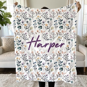 May include: A white throw blanket featuring a repeating pattern of watercolor-style flowers and leaves in shades of blue, peach, and grey. The name Harper is printed in the center in a large, dark purple, cursive script font across the fabric.