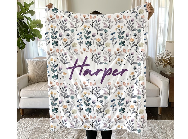 May include: A white throw blanket featuring a repeating pattern of watercolor-style flowers and leaves in shades of blue, peach, and grey. The name Harper is printed in the center in a large, dark purple, cursive script font across the fabric.