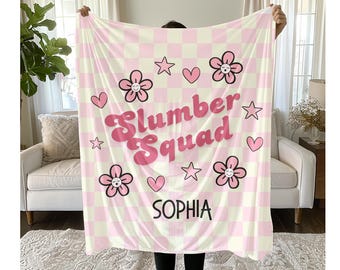 May include: A pink and white checkered blanket with the words "Slumber Squad" in pink script, along with flower and star graphics. The name "Sophia" is printed at the bottom. A cozy, decorative blanket.