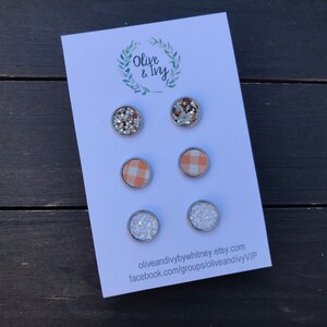 Op de afbeelding: Set van zes oorbel studs met verschillende ontwerpen. Twee oorbel studs hebben een zilveren glitterontwerp, twee oorbel studs hebben een perzikkleurig en wit ruitpatroon en twee oorbel studs hebben een wit glitterontwerp. De oorbel studs worden gepresenteerd op een witte kaart met de tekst "Olive & Ivy" en contactgegevens.