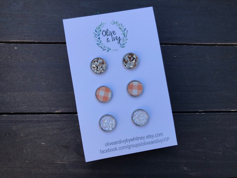 Op de afbeelding: Set van zes oorbel studs met verschillende ontwerpen. Twee oorbel studs hebben een zilveren glitterontwerp, twee oorbel studs hebben een perzikkleurig en wit ruitpatroon en twee oorbel studs hebben een wit glitterontwerp. De oorbel studs worden gepresenteerd op een witte kaart met de tekst "Olive & Ivy" en contactgegevens.