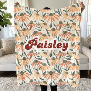 May include: A soft throw blanket featuring a repeating pattern of orange and yellow coneflower blossoms with dark green leaves on a cream background. The name Paisley is printed in the center in a bold, dark red, retro-style cursive font.