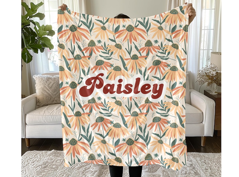 May include: A soft throw blanket featuring a repeating pattern of orange and yellow coneflower blossoms with dark green leaves on a cream background. The name Paisley is printed in the center in a bold, dark red, retro-style cursive font.