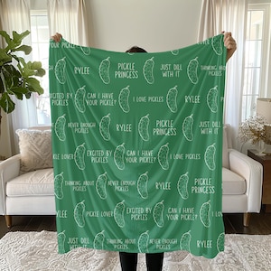 May include: A green blanket featuring white pickle illustrations and phrases such as "Pickle Princess" and "Just Dill With It." The blanket is displayed, highlighting its design and theme.