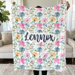May include: A rectangular throw blanket featuring a repeating watercolor floral pattern in shades of pink, blue, and yellow with green leaves. The name Lennox is printed in large, dark gray, cursive script across the center of the blanket.