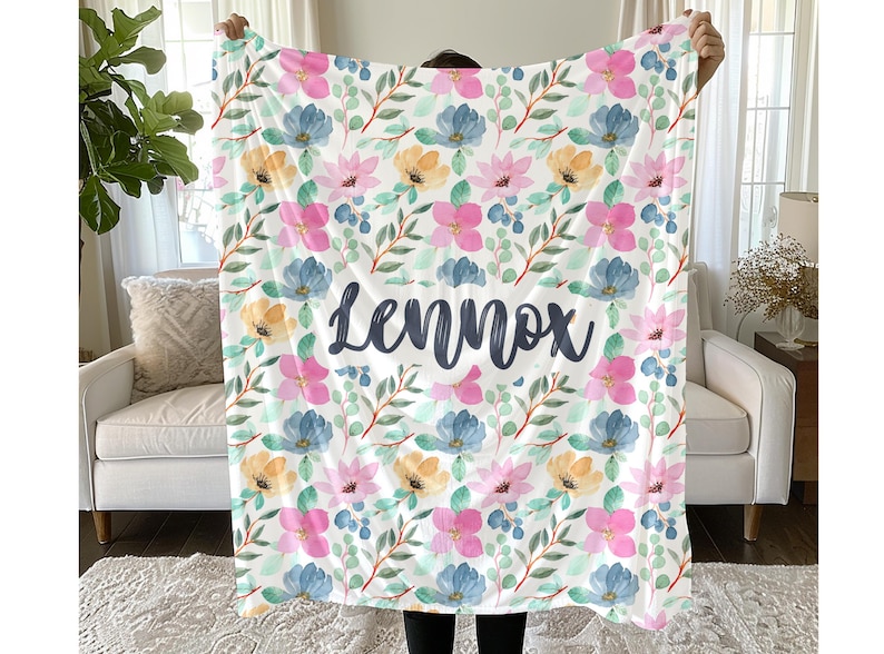 May include: A rectangular throw blanket featuring a repeating watercolor floral pattern in shades of pink, blue, and yellow with green leaves. The name Lennox is printed in large, dark gray, cursive script across the center of the blanket.
