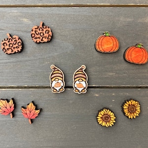 May include: A set of wooden earrings featuring various fall-themed designs, including pumpkins, gnomes, sunflowers, and leaves. The earrings are painted in shades of orange, yellow, brown, and green.