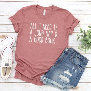 May include: A dusty rose t-shirt with the text "All I need is a long nap & a good book" in white lettering. The shirt is paired with denim shorts and white sneakers.