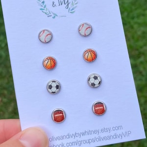 May include: A set of six silver stud earrings featuring different sports balls: baseball, basketball, football, and soccer. The earrings are displayed on a white card with the text "oliveandivybywhitney.etsy.com" and "facebook.com/groups/oliveandivyVIP".