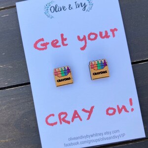 K&ouml;nnte beinhalten: Ein Paar Ohrringe aus Holz in Form einer Schachtel mit Buntstiften. Die Schachtel ist braun mit dem aufgedruckten Wort "Crayons". Die Buntstift sind bunt und in einer Reihe angeordnet. Die Ohrringe befinden sich auf einer wei&szlig;en Karte mit dem Text "Get your Cray on!" und der Webadresse des Shops.