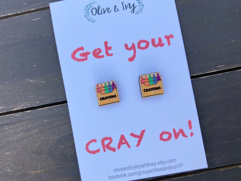 K&ouml;nnte beinhalten: Ein Paar Ohrringe aus Holz in Form einer Schachtel mit Buntstiften. Die Schachtel ist braun mit dem aufgedruckten Wort "Crayons". Die Buntstift sind bunt und in einer Reihe angeordnet. Die Ohrringe befinden sich auf einer wei&szlig;en Karte mit dem Text "Get your Cray on!" und der Webadresse des Shops.